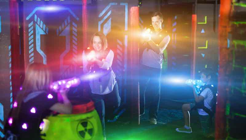Excited kids and theirs parents in bright beams of laser guns during laser tag game in dark room
