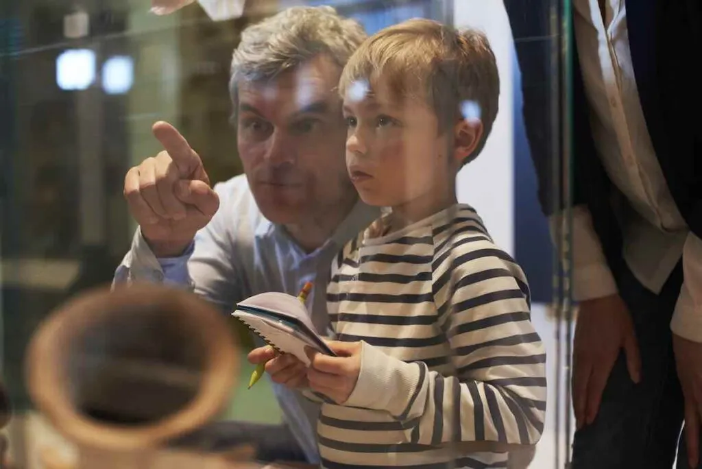 Father And Son Look At Artifacts In Case On Trip To Museum