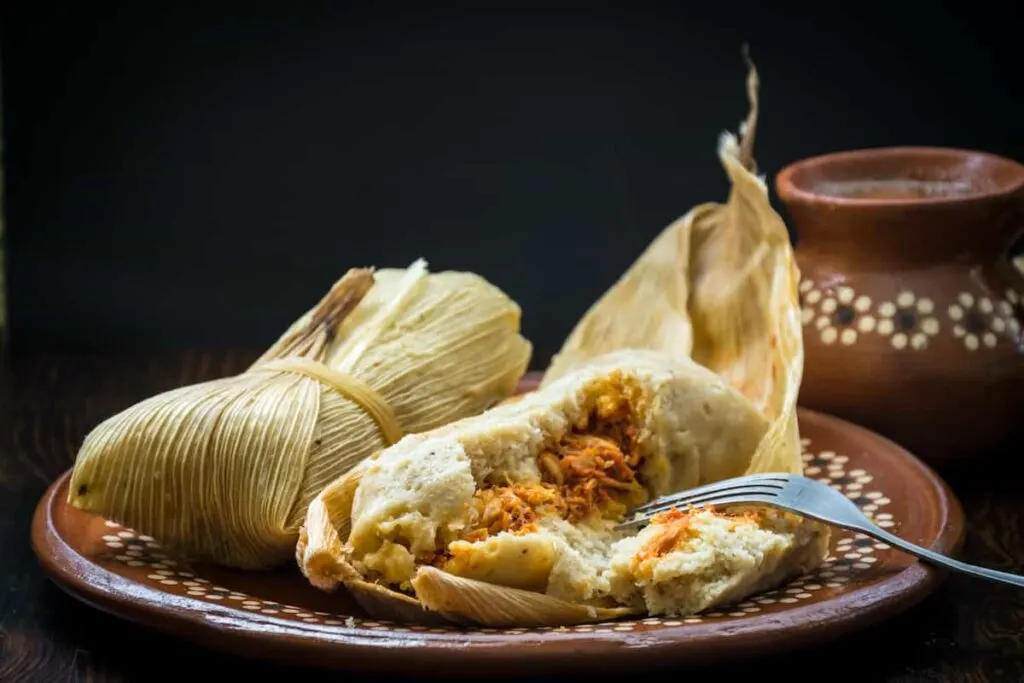 A tamale still wrapped in a corn husk and another one cut into with a fork to reveal the pork filling.